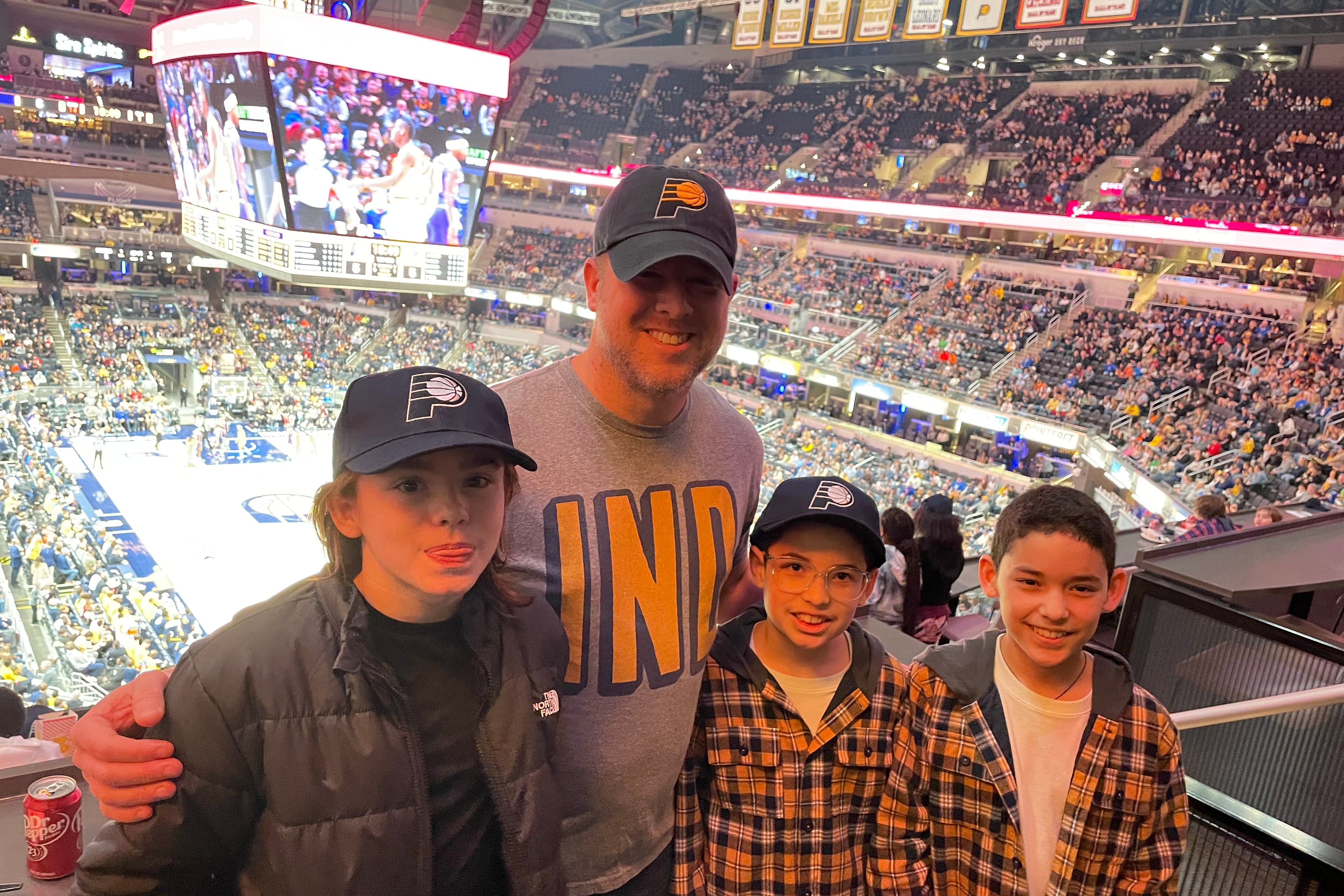 A man in a Pacers basketball hat stands smiling with three other students with a Pacers basketball game going on in the background.