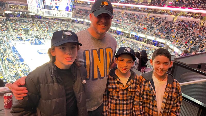 A man in a Pacers basketball hat stands smiling with three other students with a Pacers basketball game going on in the background.