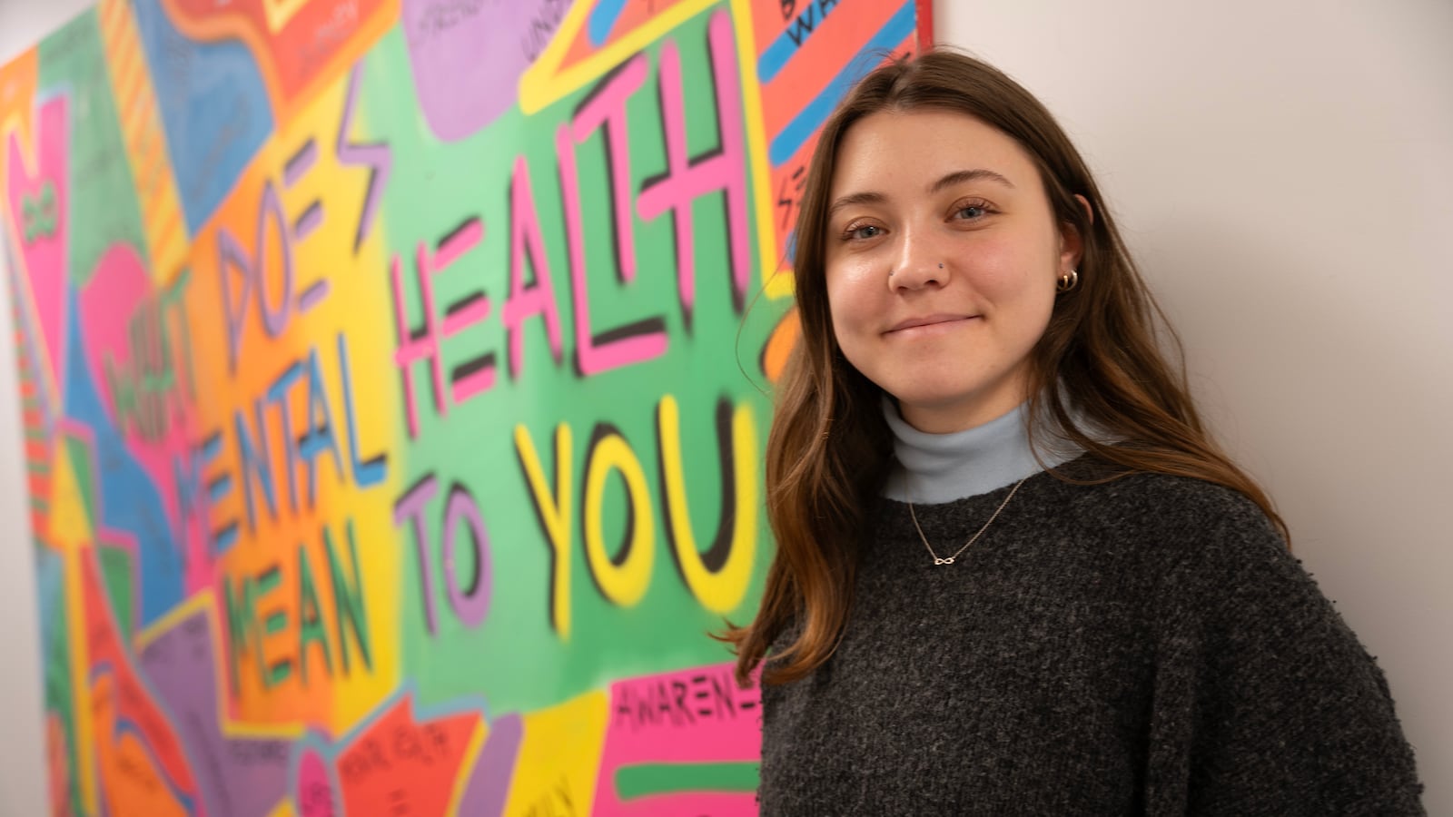 A young white student with long dark hair stands next to a colorful painting that is hanging on a white wall.