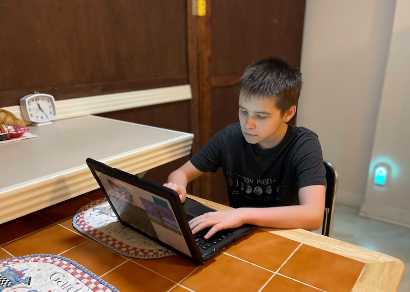 A young boy wearing a black t-shirt, sits at a table working on his computer in his home.