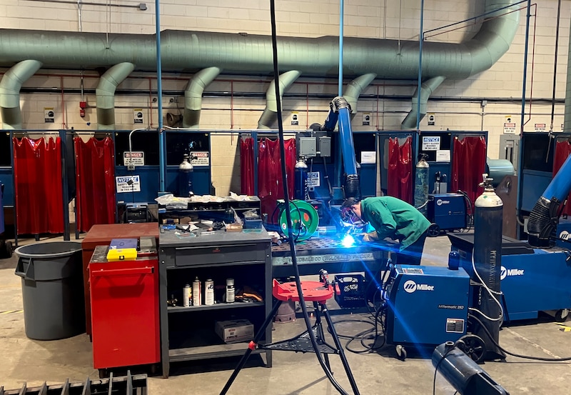 A student welds in the background of a large welding room of a high school.