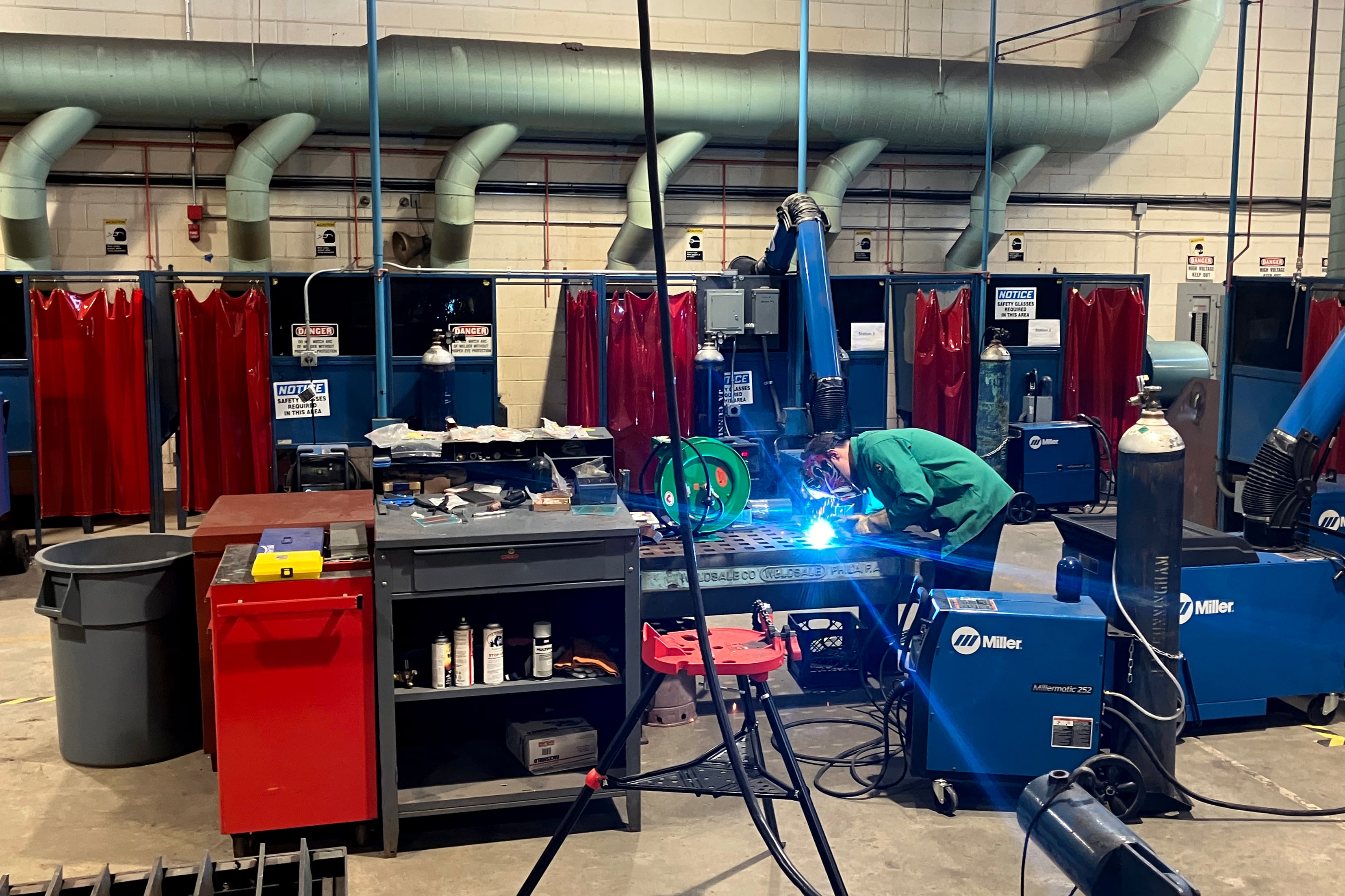 A student welds in the background of a large welding room of a high school.