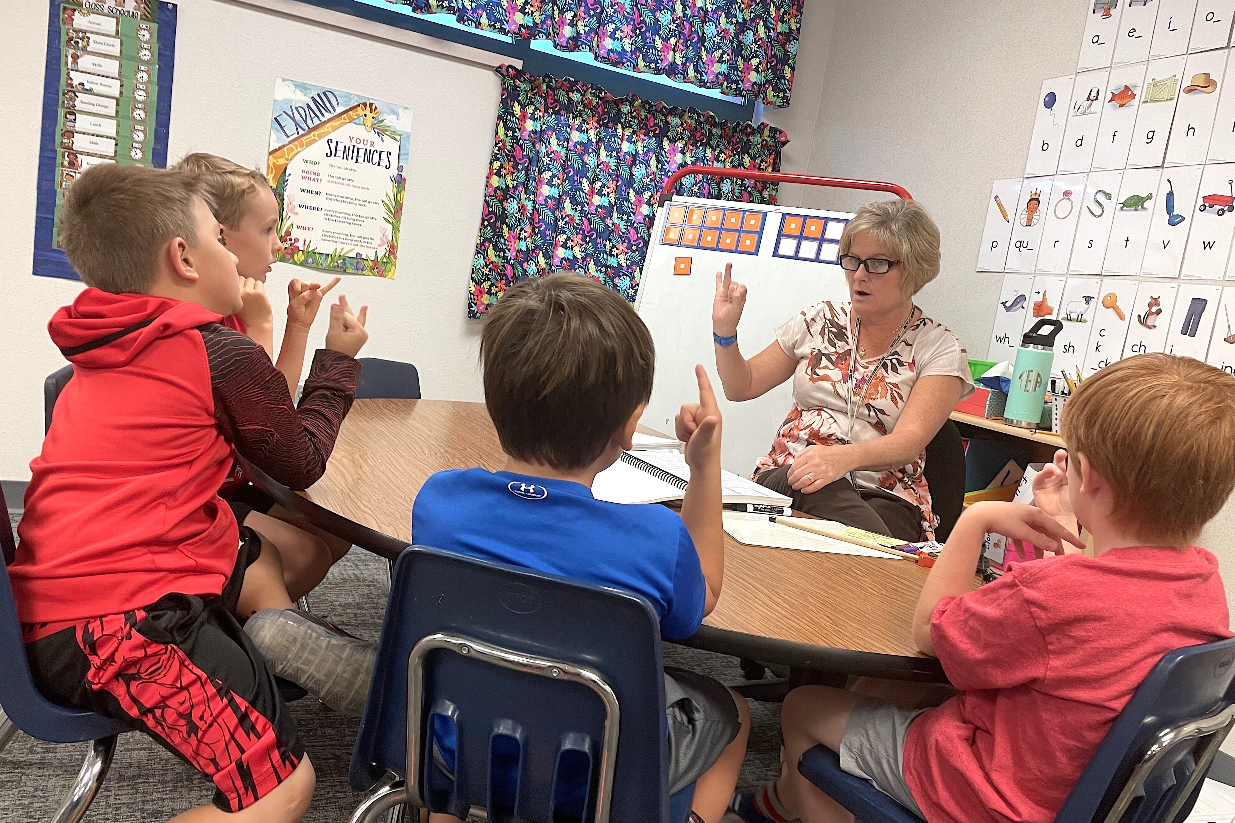 A woman works with four of her students on a reading lesson at a small table.