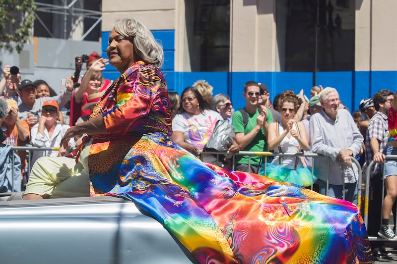 A Black woman with gray hair wears a rainbow coat as she rides in a convertible through a Pride parade. Onlookers cheer.