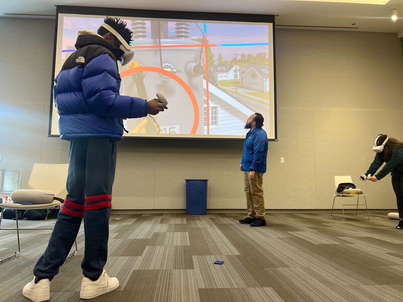 Three people stand in a room near a large screen and two people are wearing virtual reality goggles.