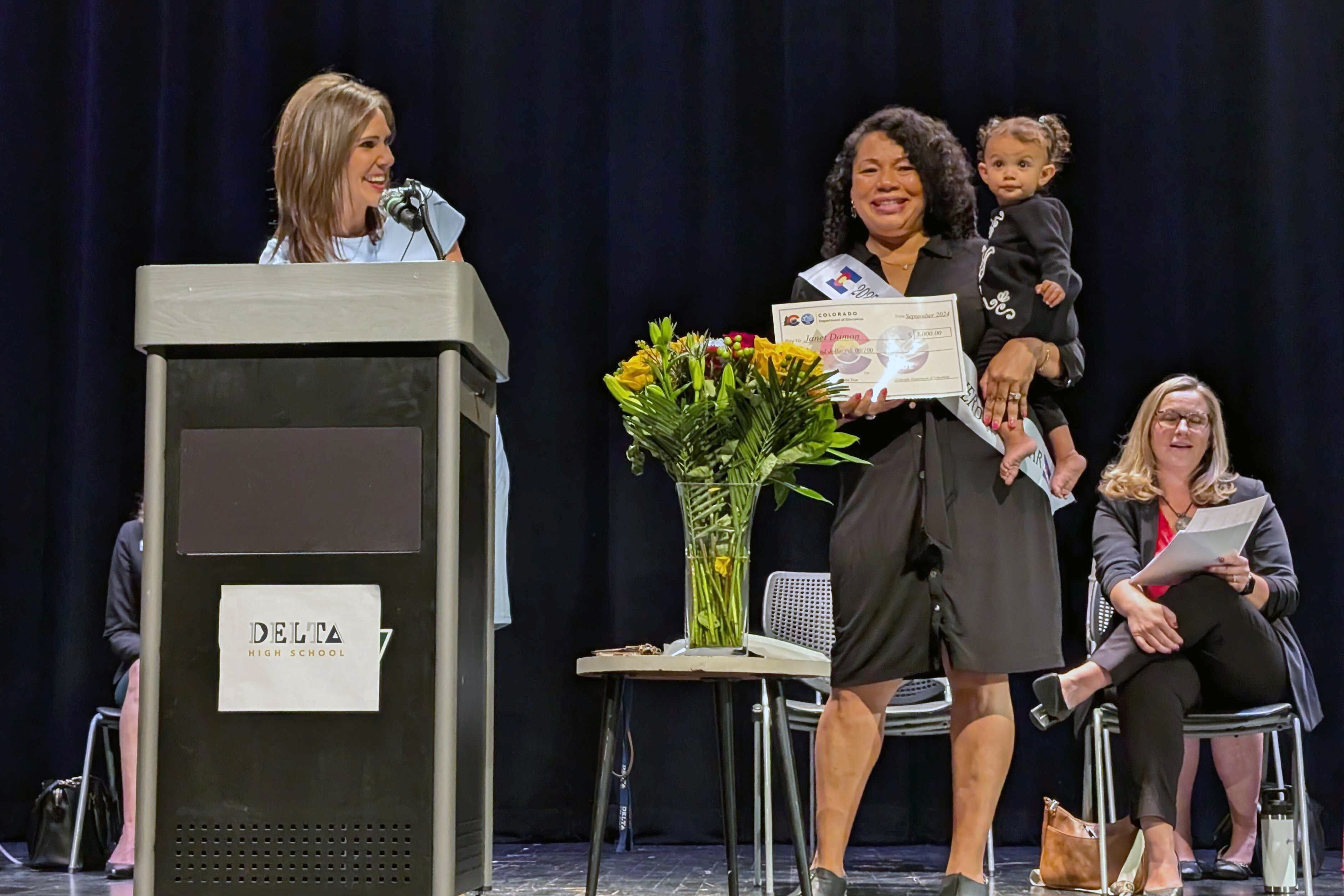 A woman wearing a dark dress and holding a child and an award certificate stands next to a woman who is behind a podium on a stage.