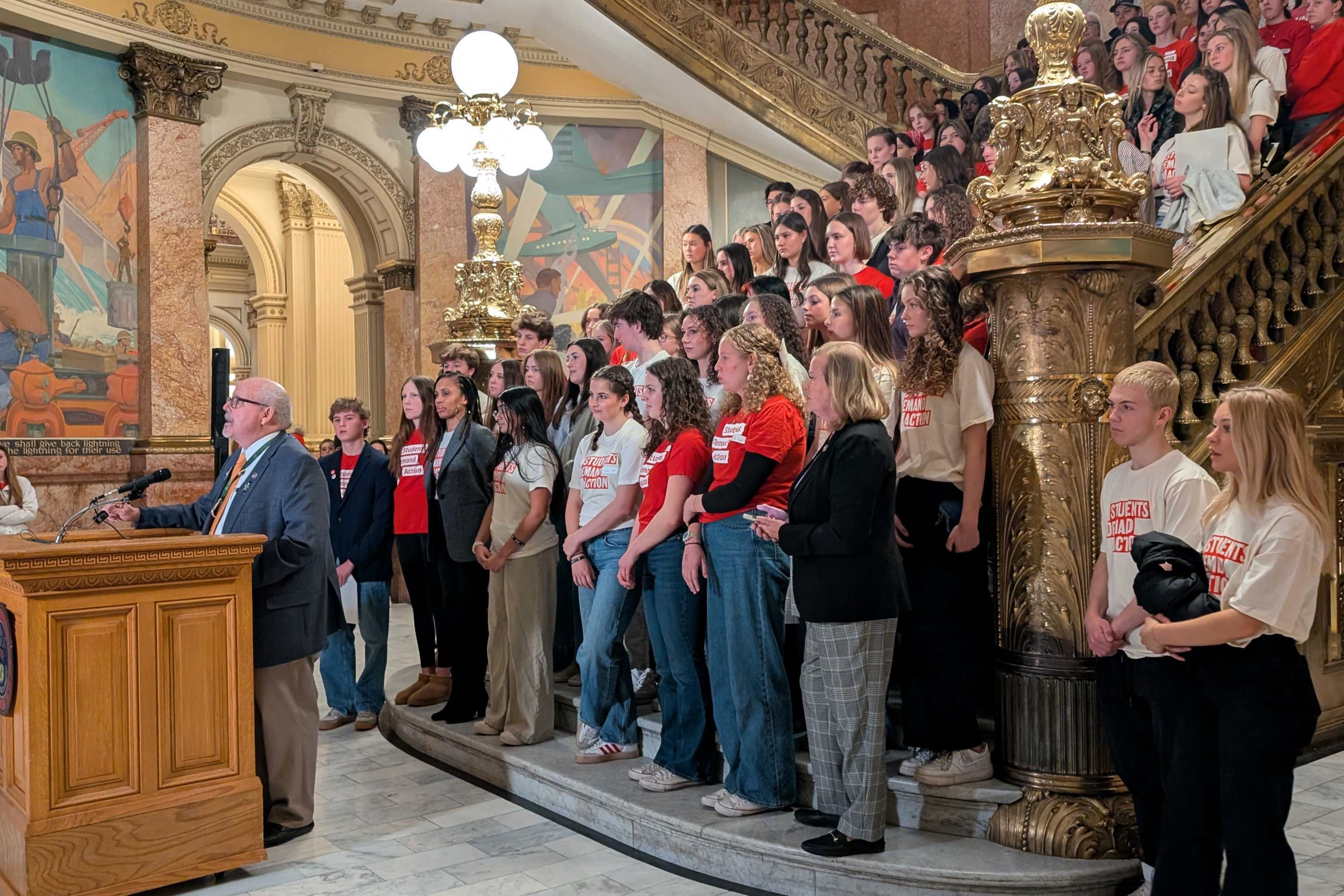 A large group of high school students and some adults and most of them are wearing a read shirt all stand on the stairs of a capitol building.