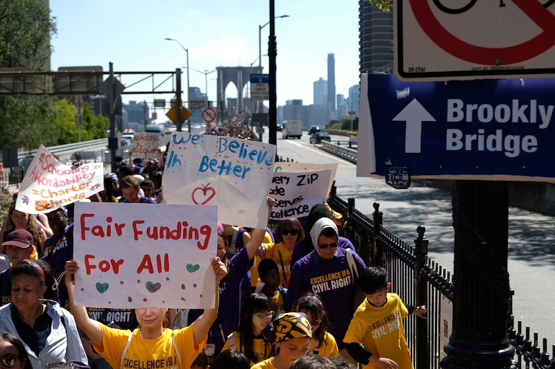 A giant crowd of people, some holding signs and some wearing yellow shirts walk in a line with the Brooklyn bridge in the background.