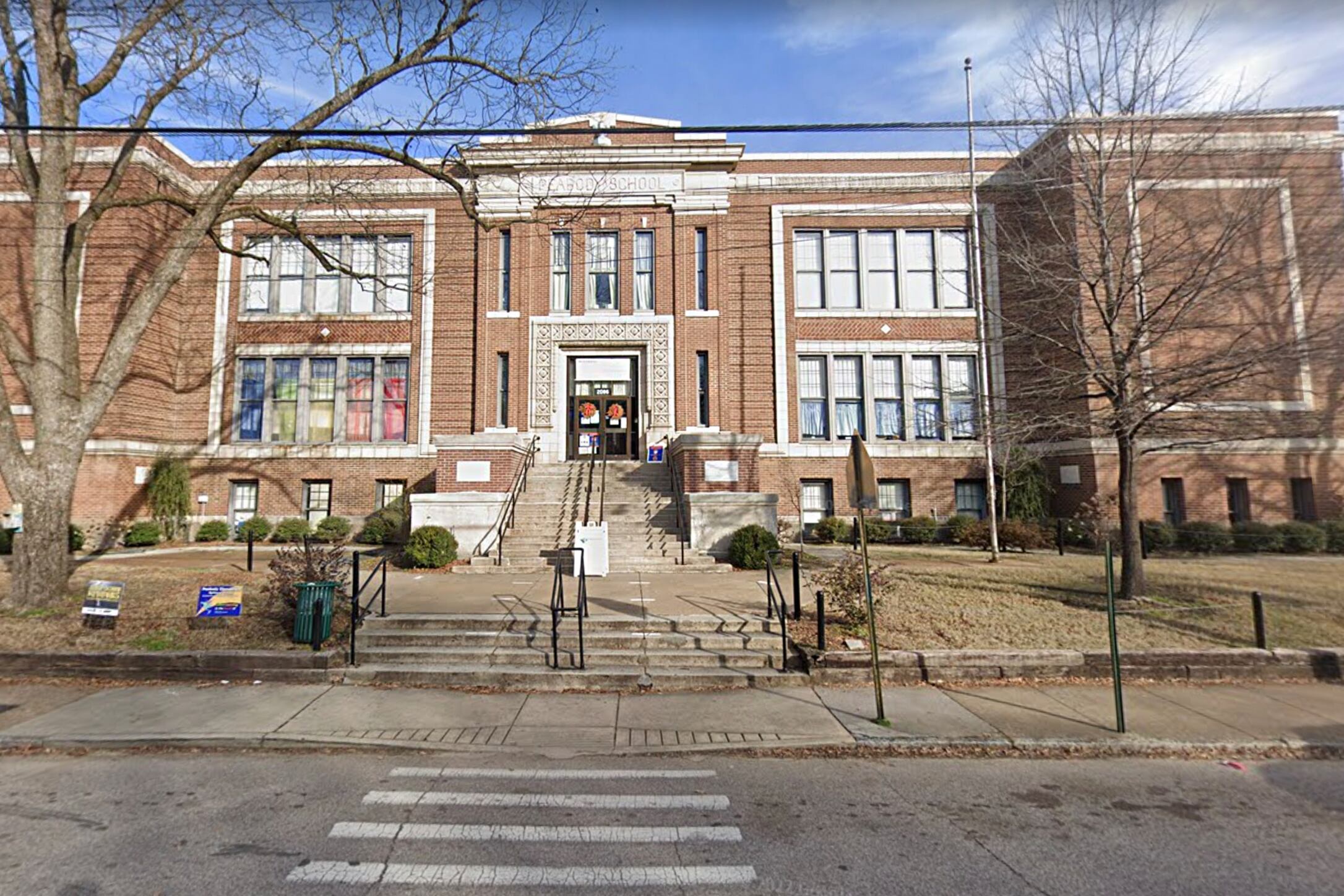 The exterior of a two-story brick school building, with steps leading up to a doorway, viewed from across the street.