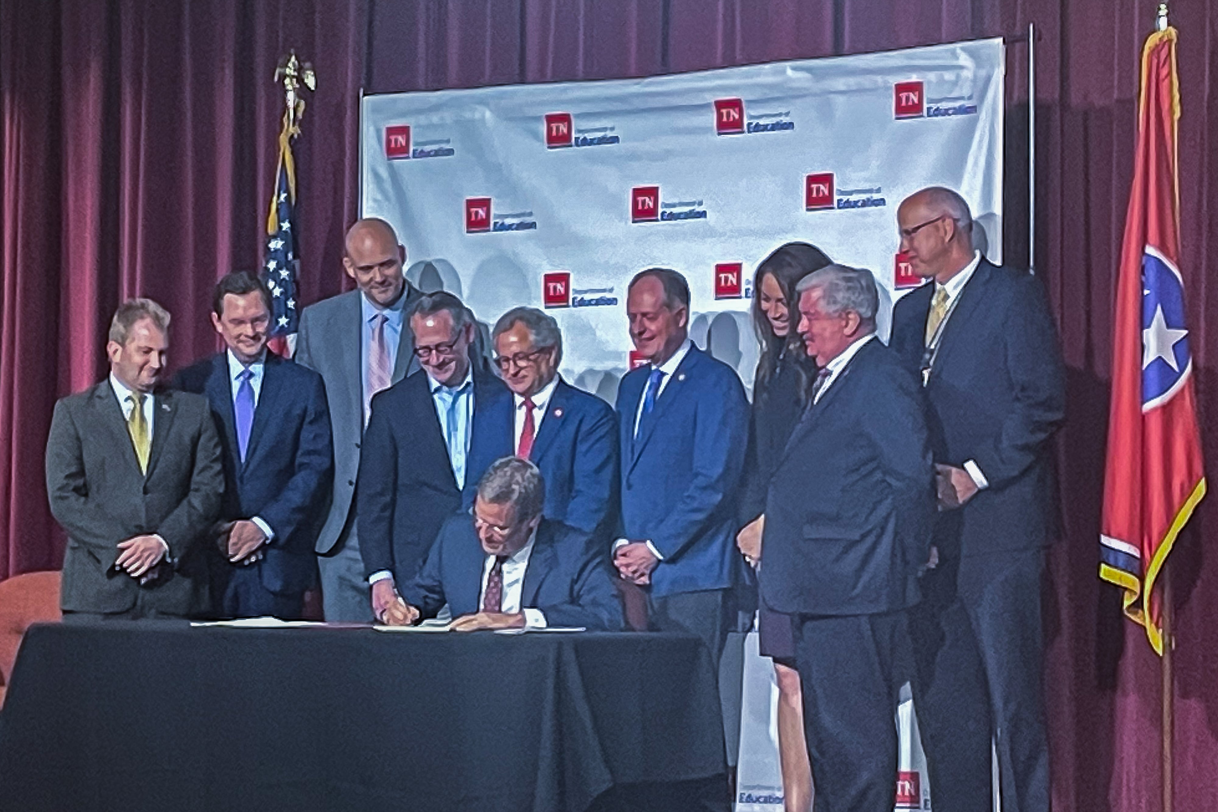 A man wearing a suit signs a paper on a desk while a group of men and one woman stand smiling behind him.