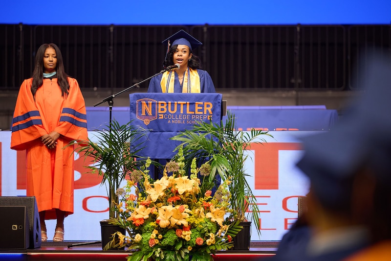 A high school senior with dark hair and wearing a blue and yellow cap and gown speaks during a graduation ceremony behind a podium that reads "Butler" while an adult wearing an orange gown stands to the right.