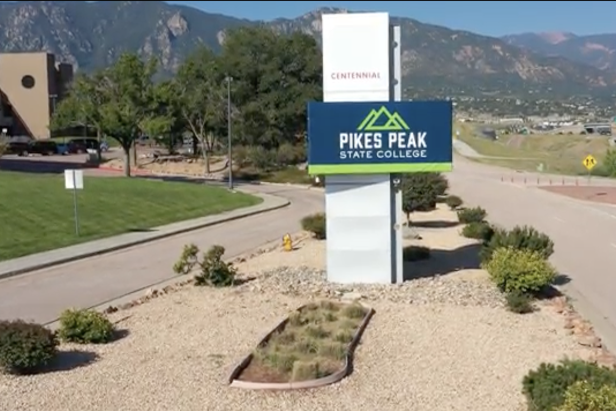 A blue and green sign is attached to a white pillar in the median of a street.