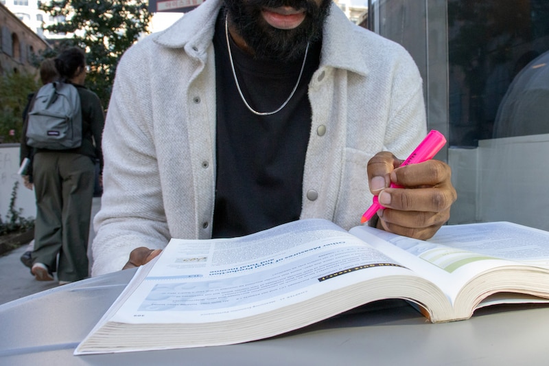 A person holds a pink highlighter while reading through a textbook.