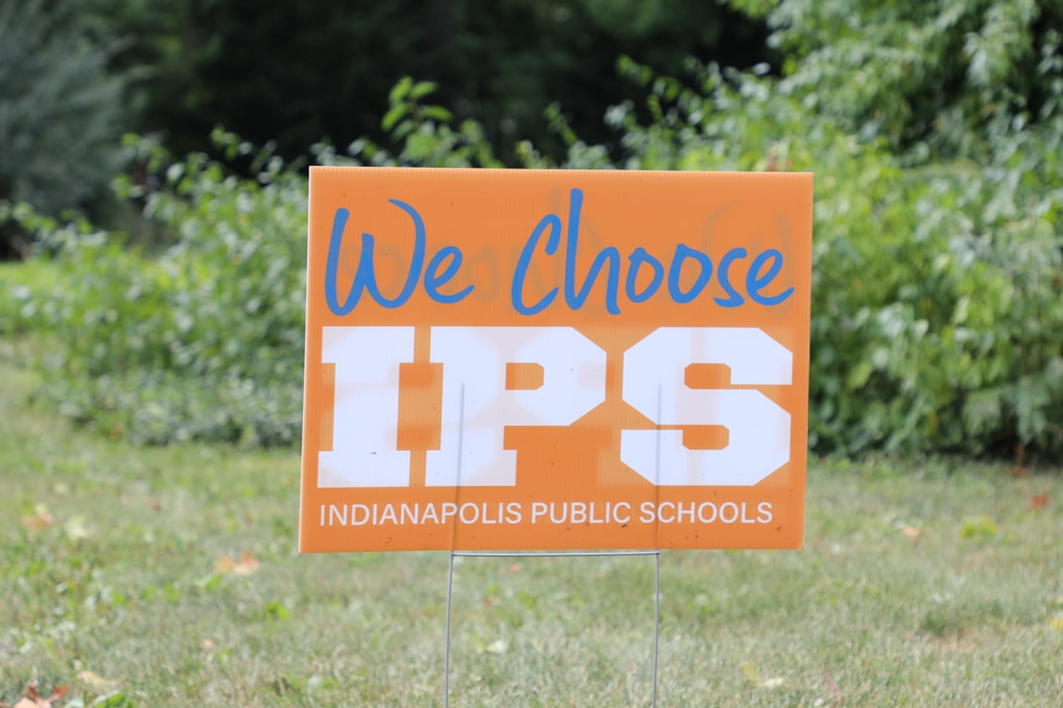 An orange lawn sign says “We Choose IPS Indianapolis Public Schools” stands on green grass. 