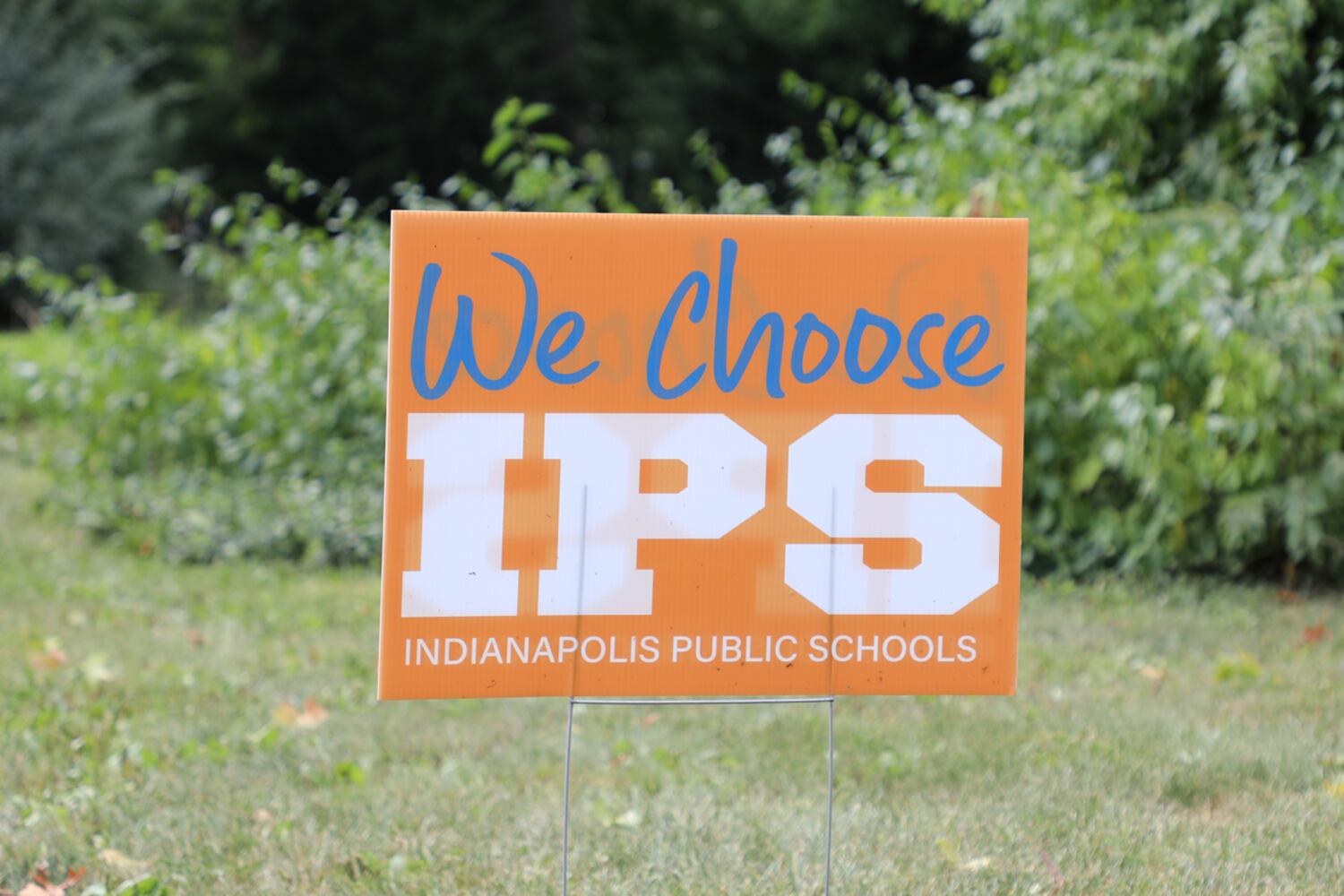 An orange lawn sign says “We Choose IPS Indianapolis Public Schools” stands on green grass.