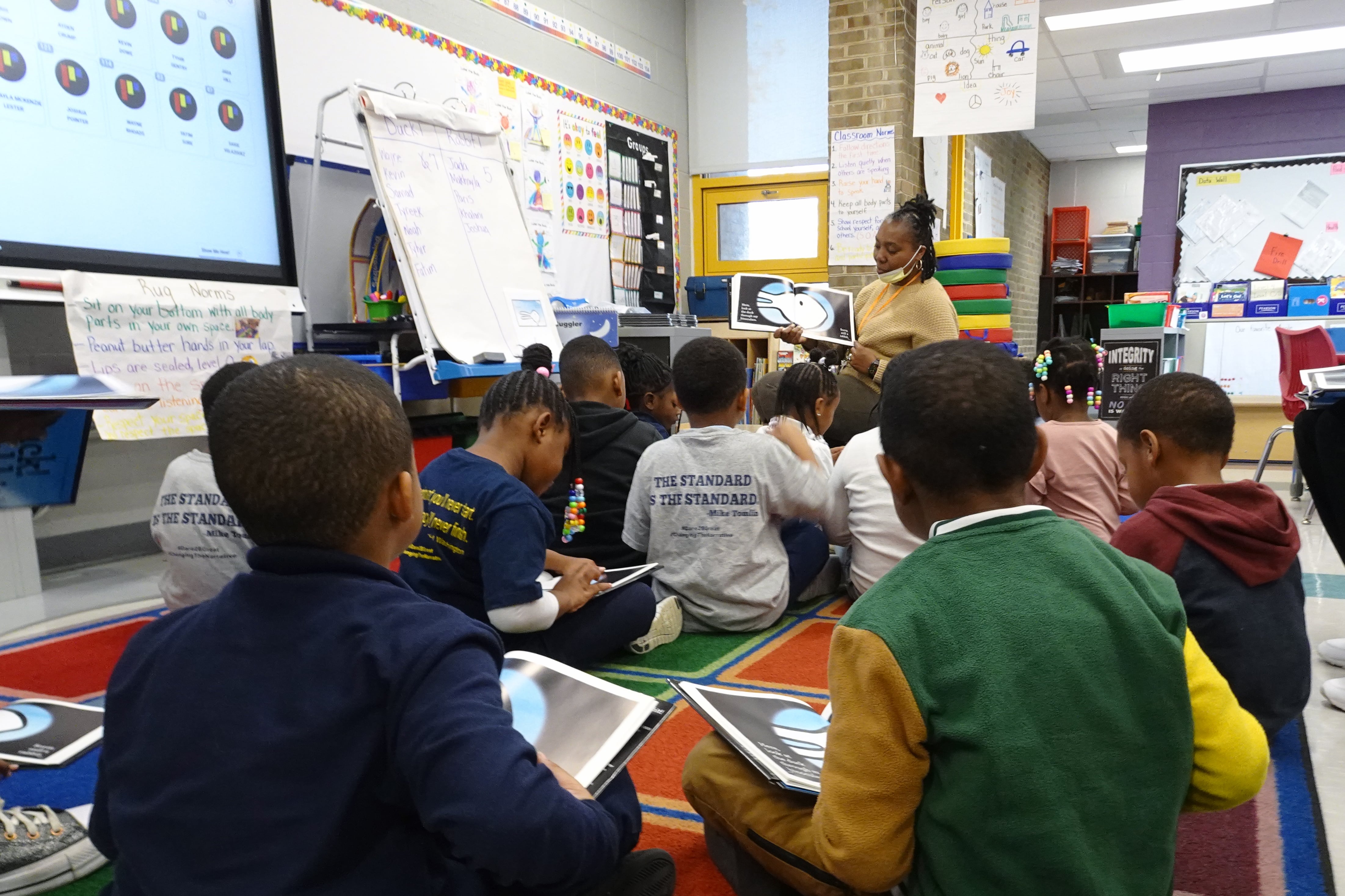 A large group of young students sit on the floor reading a book while a teacher wearing a bright yellow top reads to the students.