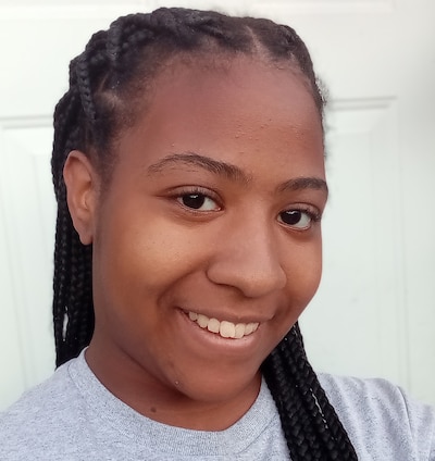A high school student with long dark braids poses for a photograph in front of a white door.