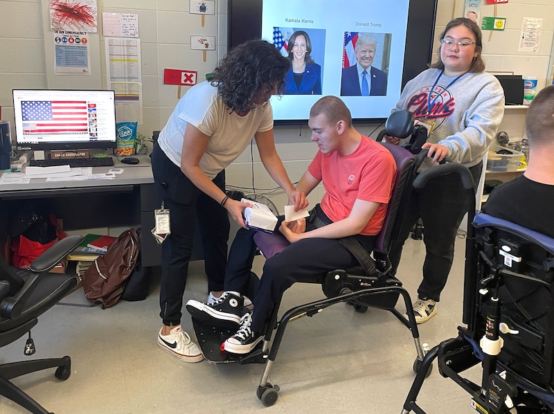 Two people stand around a student in a chair in a classroom.