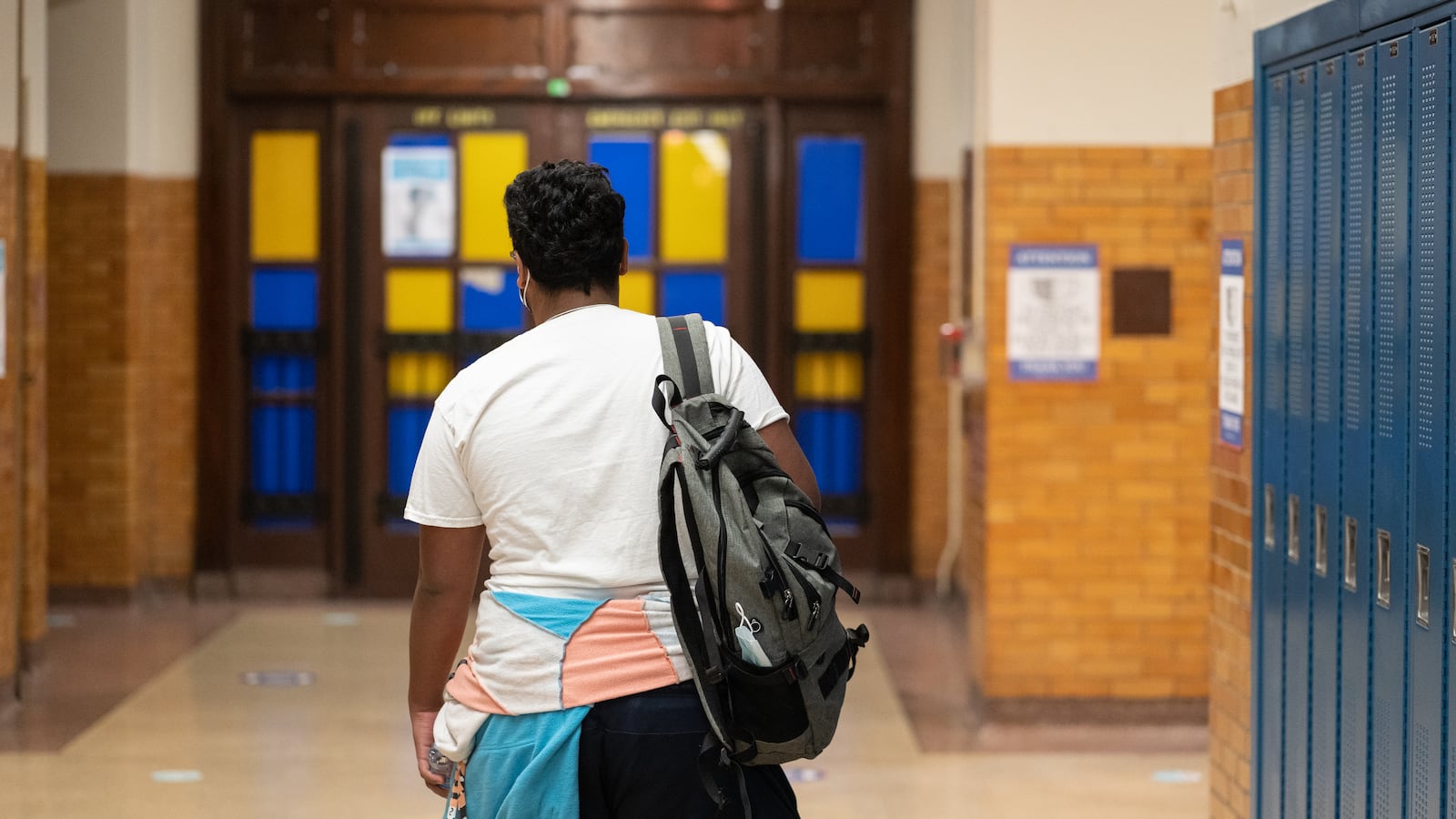 A young man walks through a school hallways, wearing a backpack, white shirt, and blue, pink, and white windbreaker tied around his waist.