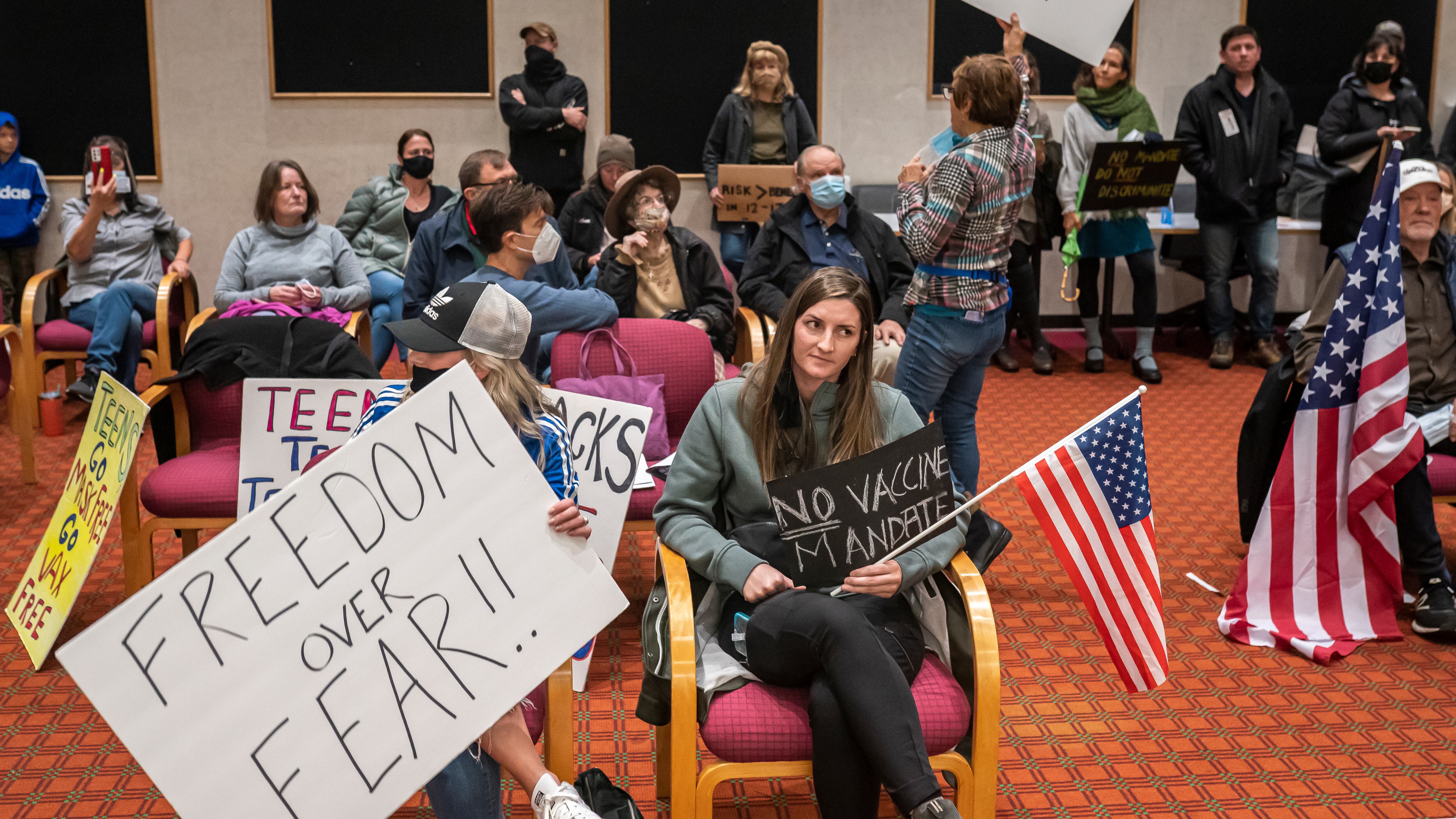 Several citizens hold American flags and signage while protesting at a school board meeting. The sign closes to the front of the crowd reads, “FREEDOM OVER FEAR” in large block letters.