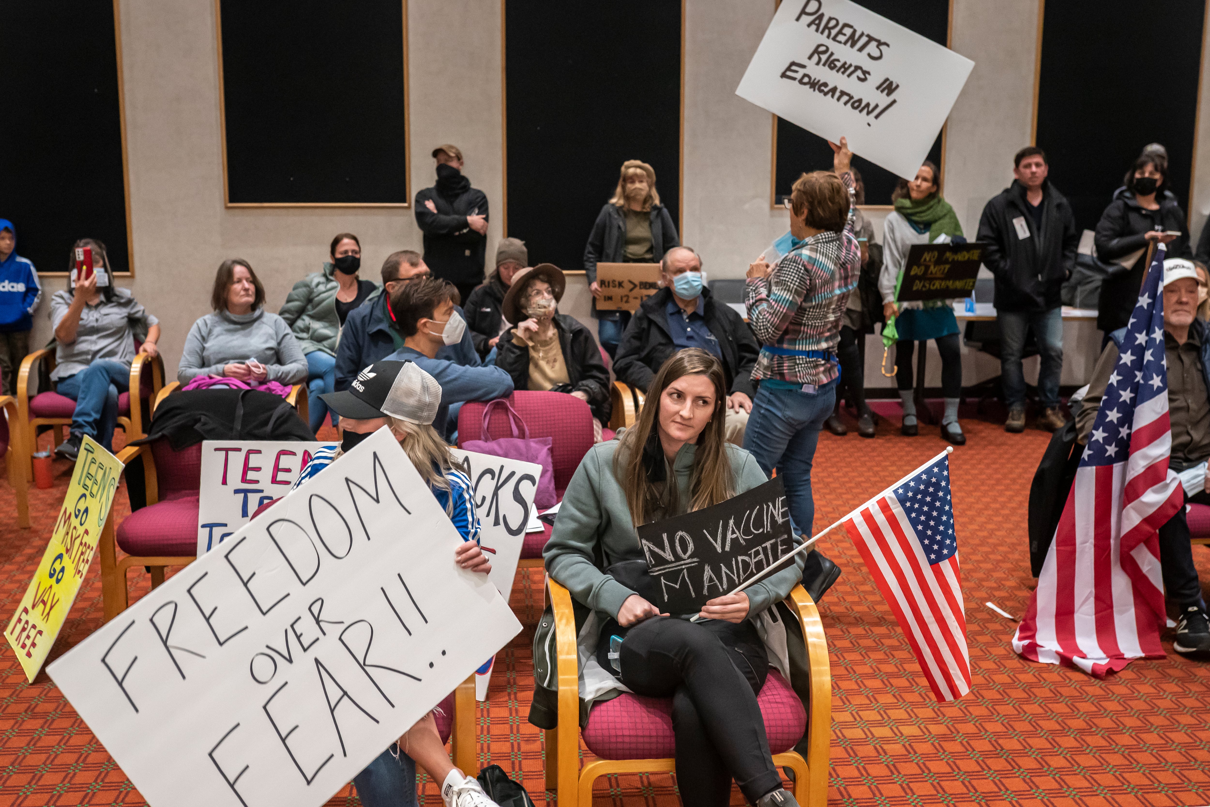 Several citizens hold American flags and signage while protesting at a school board meeting. The sign closes to the front of the crowd reads, “FREEDOM OVER FEAR” in large block letters.