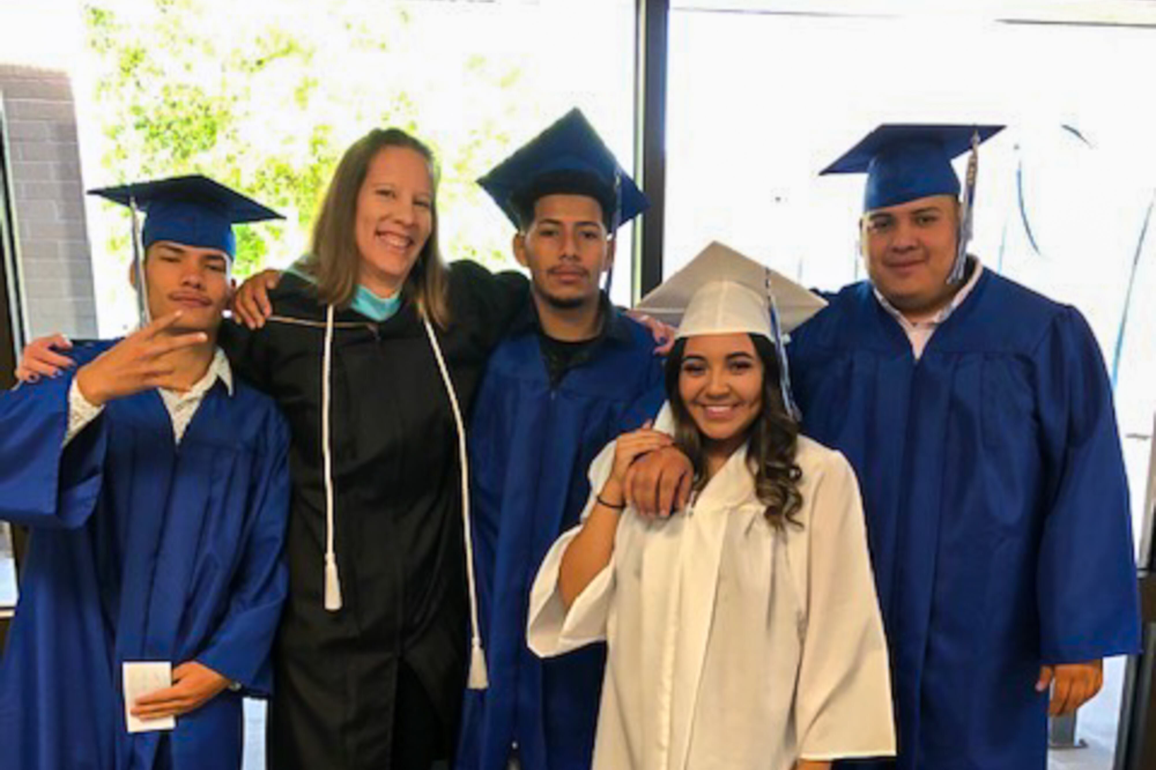 A teacher stands with four graduating seniors who are wearing blue and white graduation gowns.