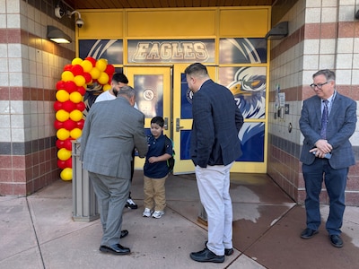 Four men in suits stand around a young student outside of a school building entrance.
