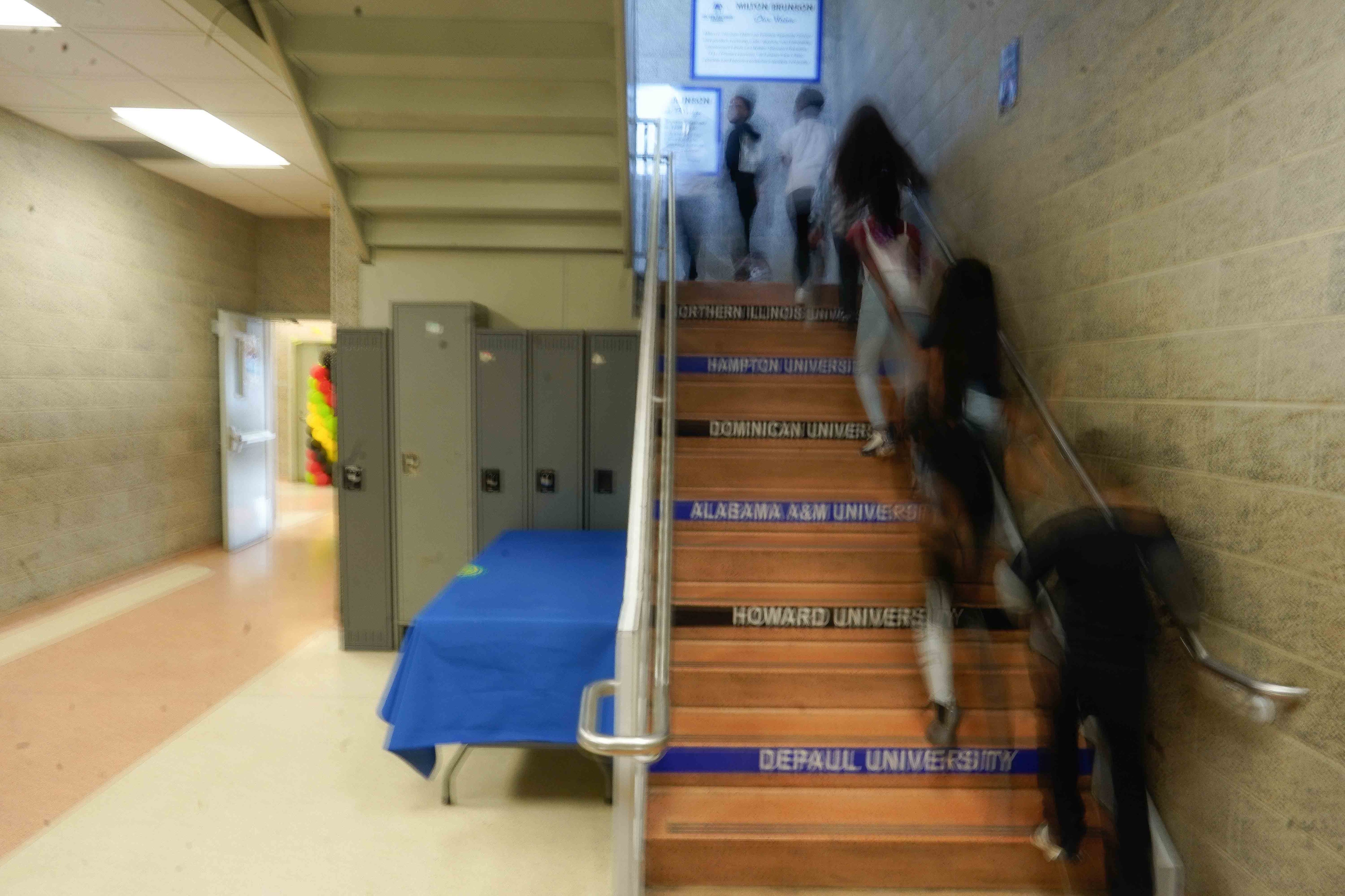 Students walk up stairs with the names of colleges on the front. There are lockers and a hallway to the left of the stairs.