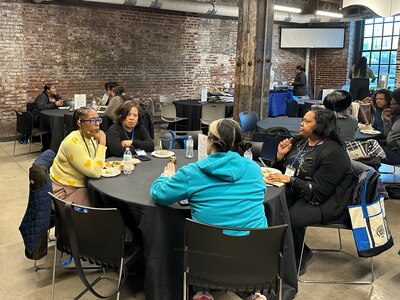 A group of adults sit at a round table with a brick wall and other tables in the background.