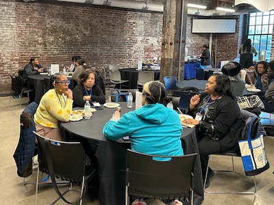 A group of adults sit at a round table with a brick wall and other tables in the background.