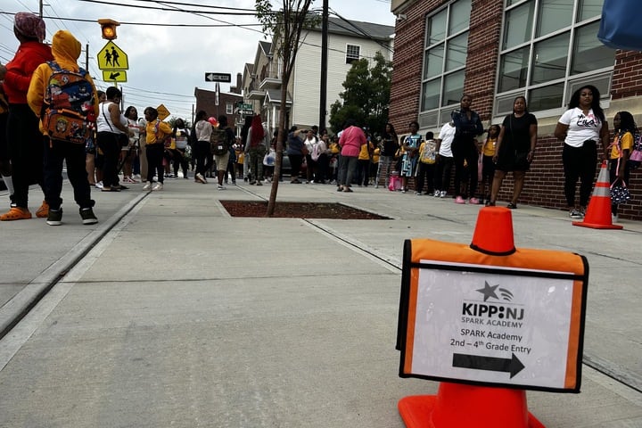 An orange cone holds a sign that says KIPP NJ as parents and children line up against a brick building.