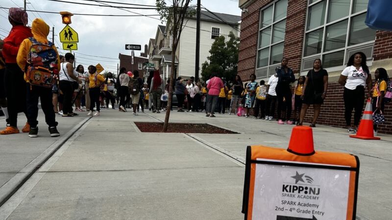 An orange cone holds a sign that says KIPP NJ as parents and children line up against a brick building.