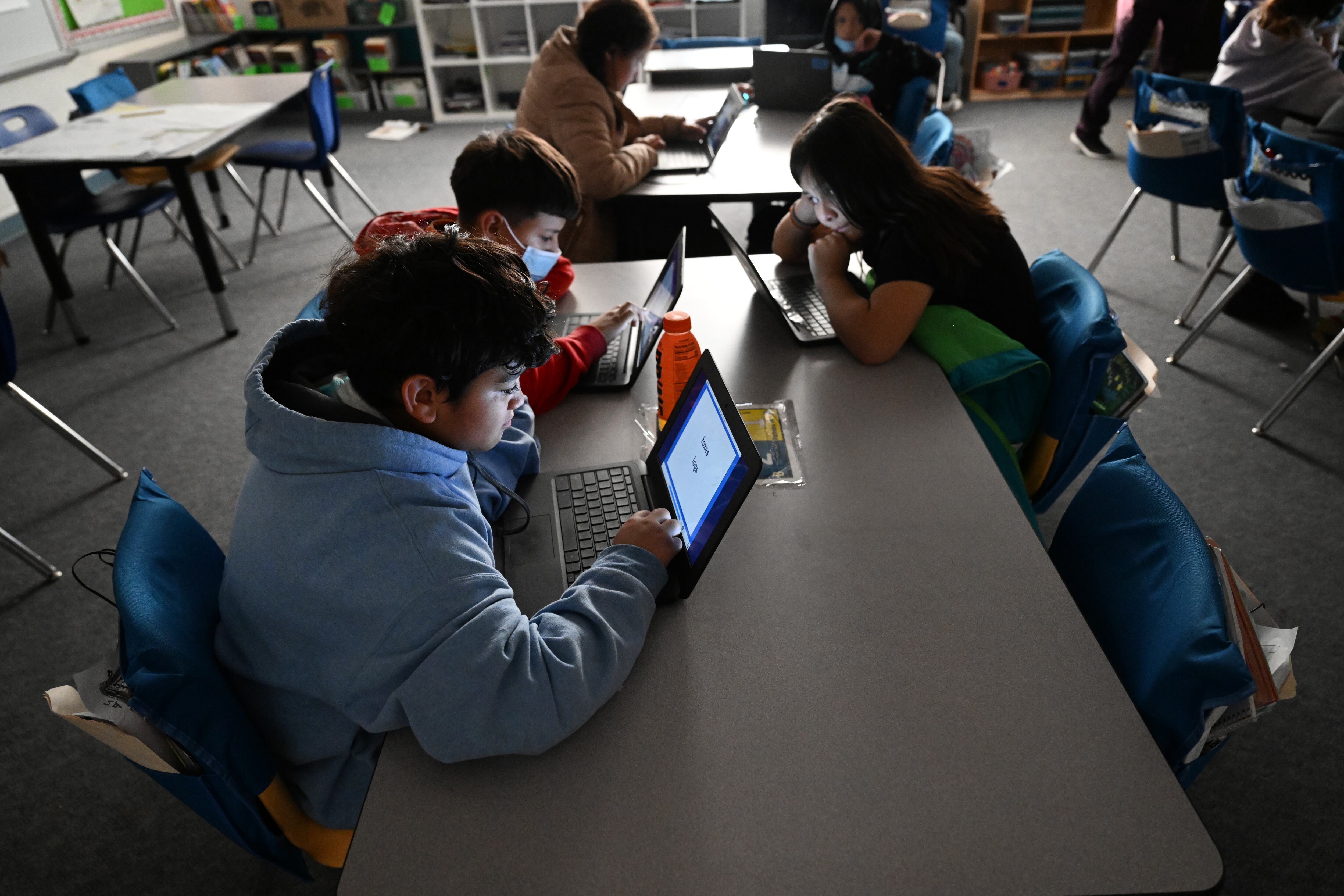 Students work on laptops in a darkened room. They’re sitting at long tables. One boy’s face is lit up by the glow of his screen.