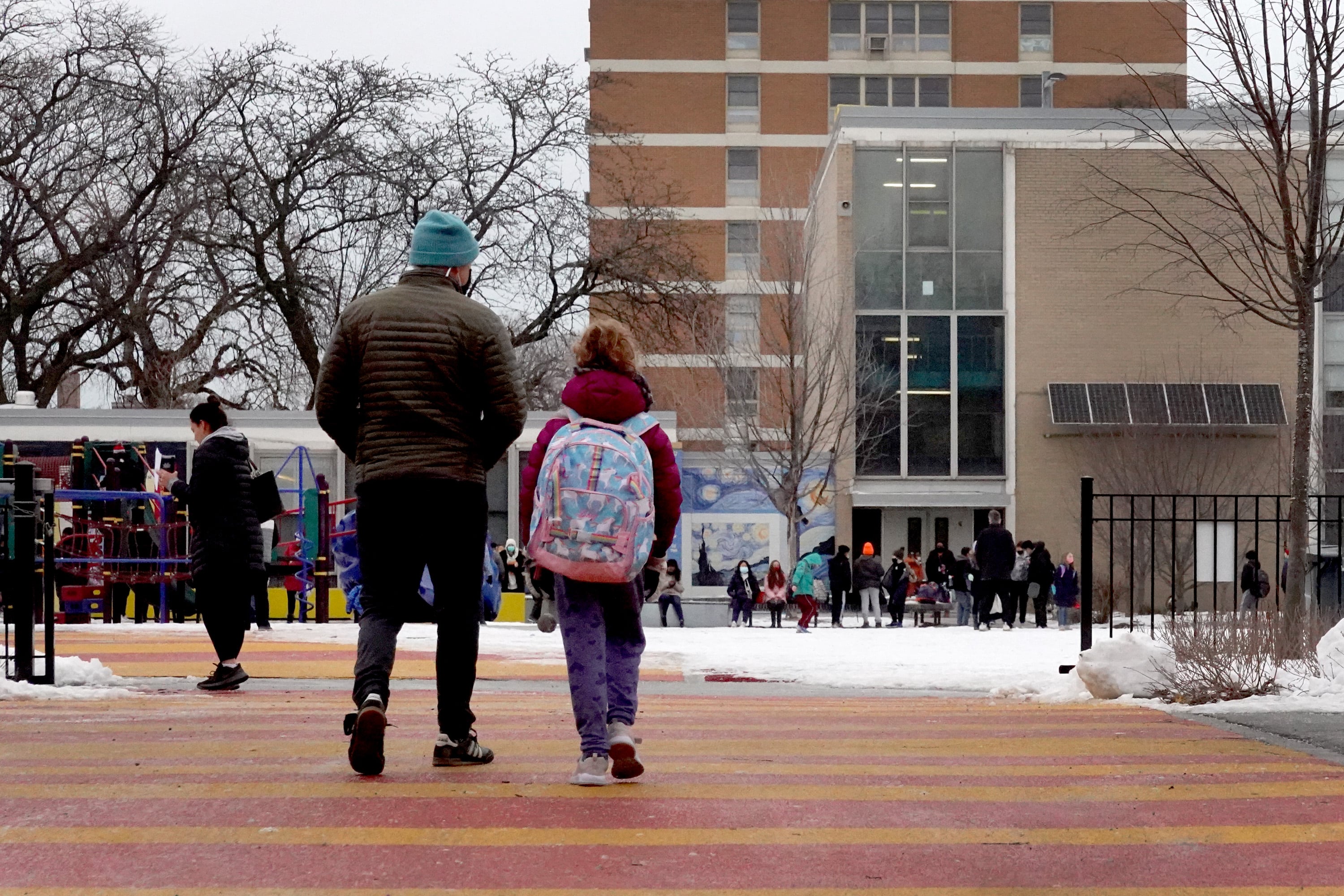 A student, wearing a knit cap and carrying a backpack, walks to a brick school building with an adult.