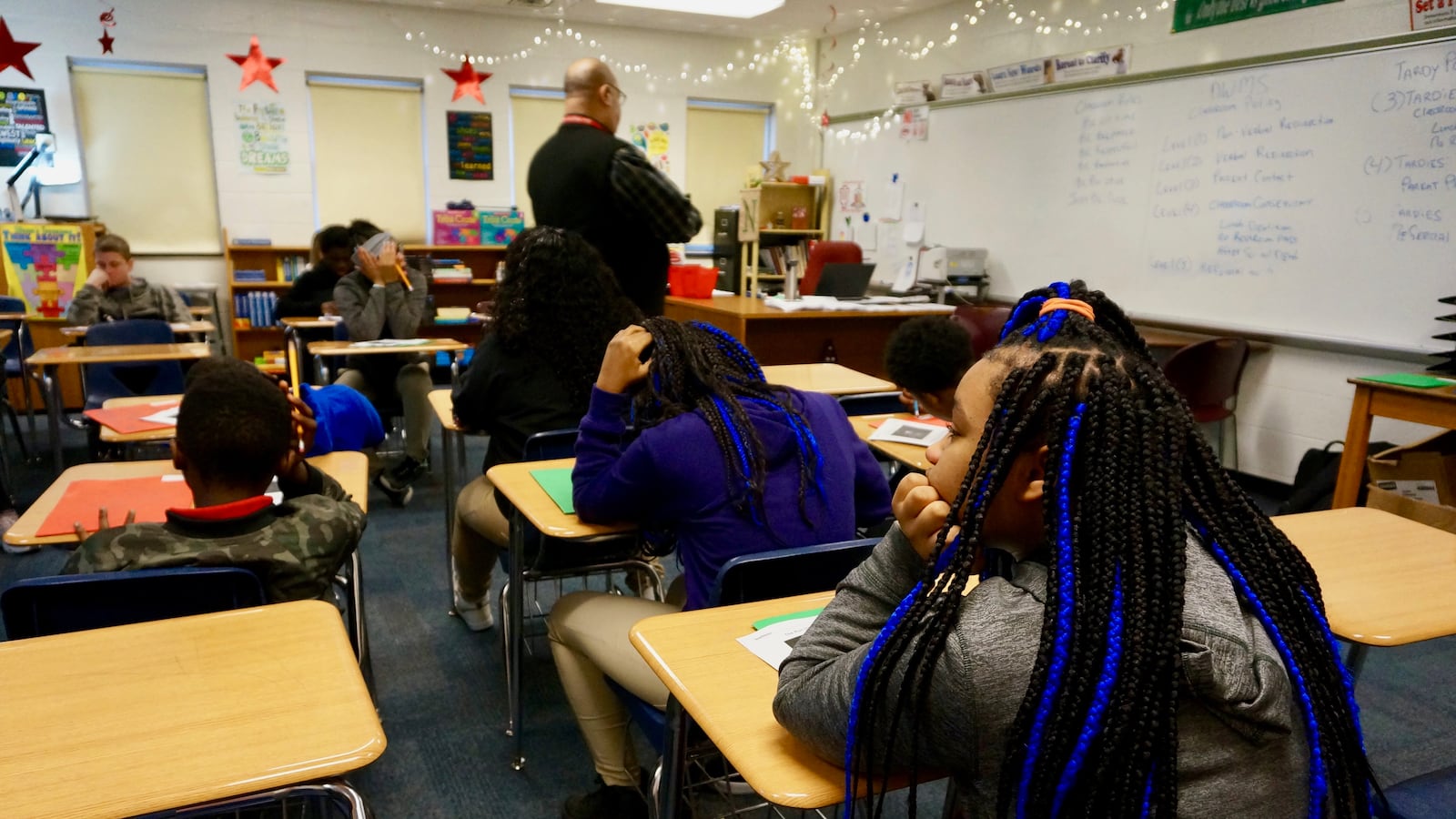 Three students in the foreground sit at desks in a classroom with their backs to the camera. A teacher walks through a line of desks in the background.