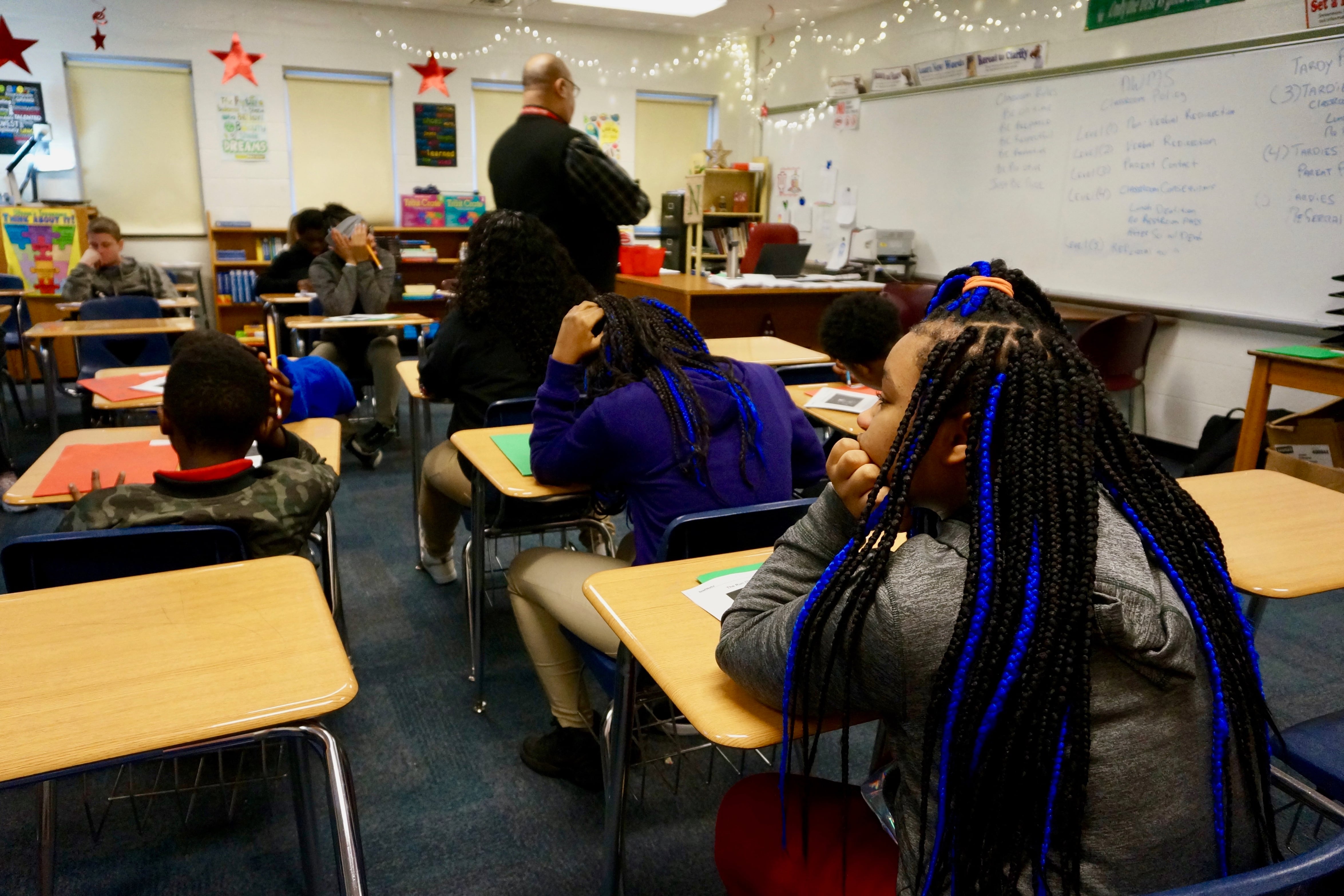 Three students in the foreground sit at desks in a classroom with their backs to the camera. A teacher walks through a line of desks in the background.