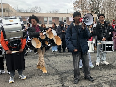 A photograph of a student talking into a loud speaker with a group of students holding protest signs and percussionists.