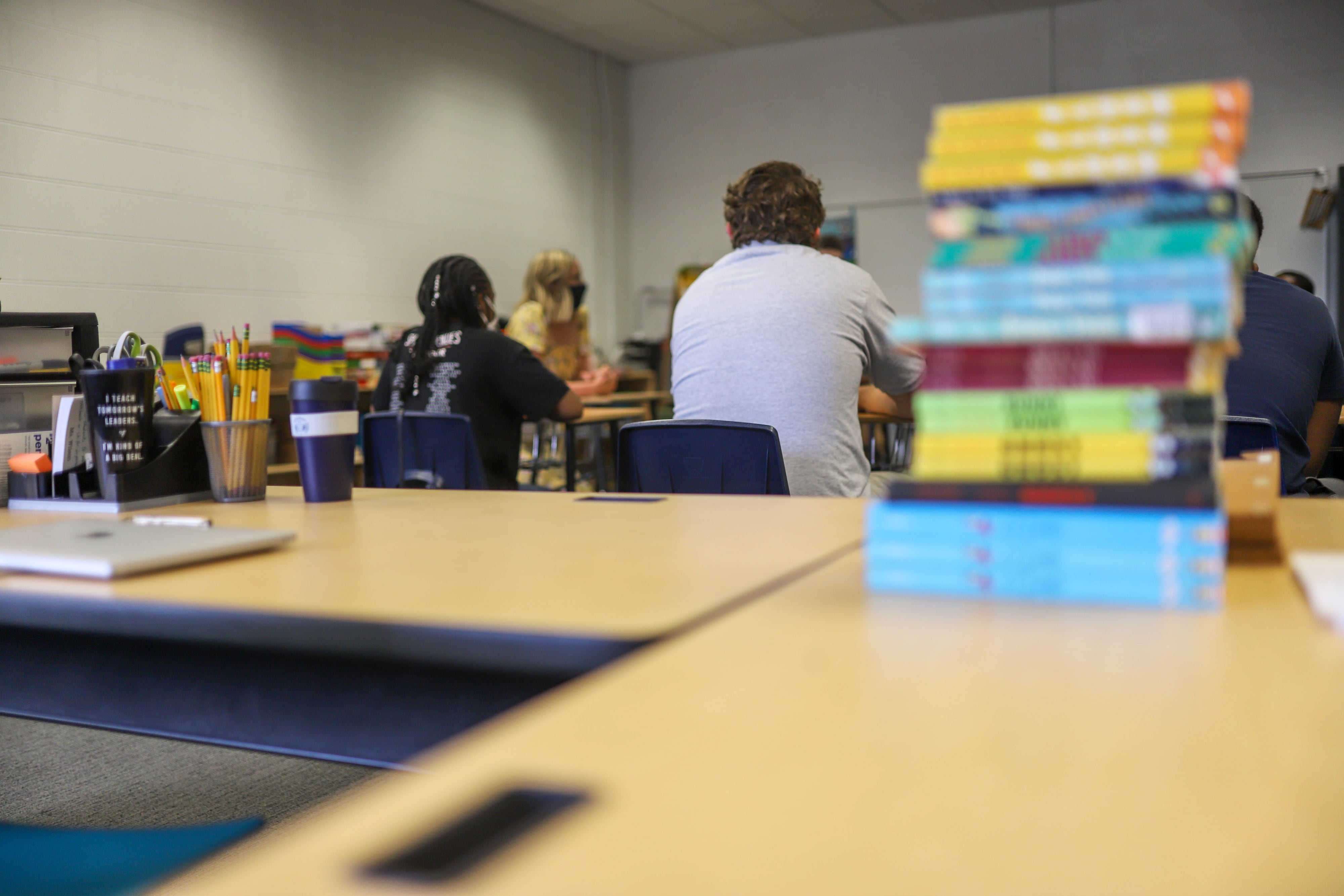 A stack of books on top of a desk is in the foreground while in the background are students sitting facing a board.