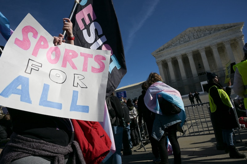 A photograph of advocates holding banners and signs rally outside the US Supreme Court, a white building with pillars.
