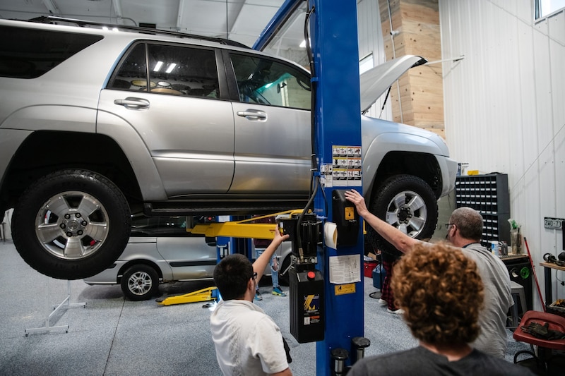 Two men hold a blue pole while another man looks on in front of a car on a lift.