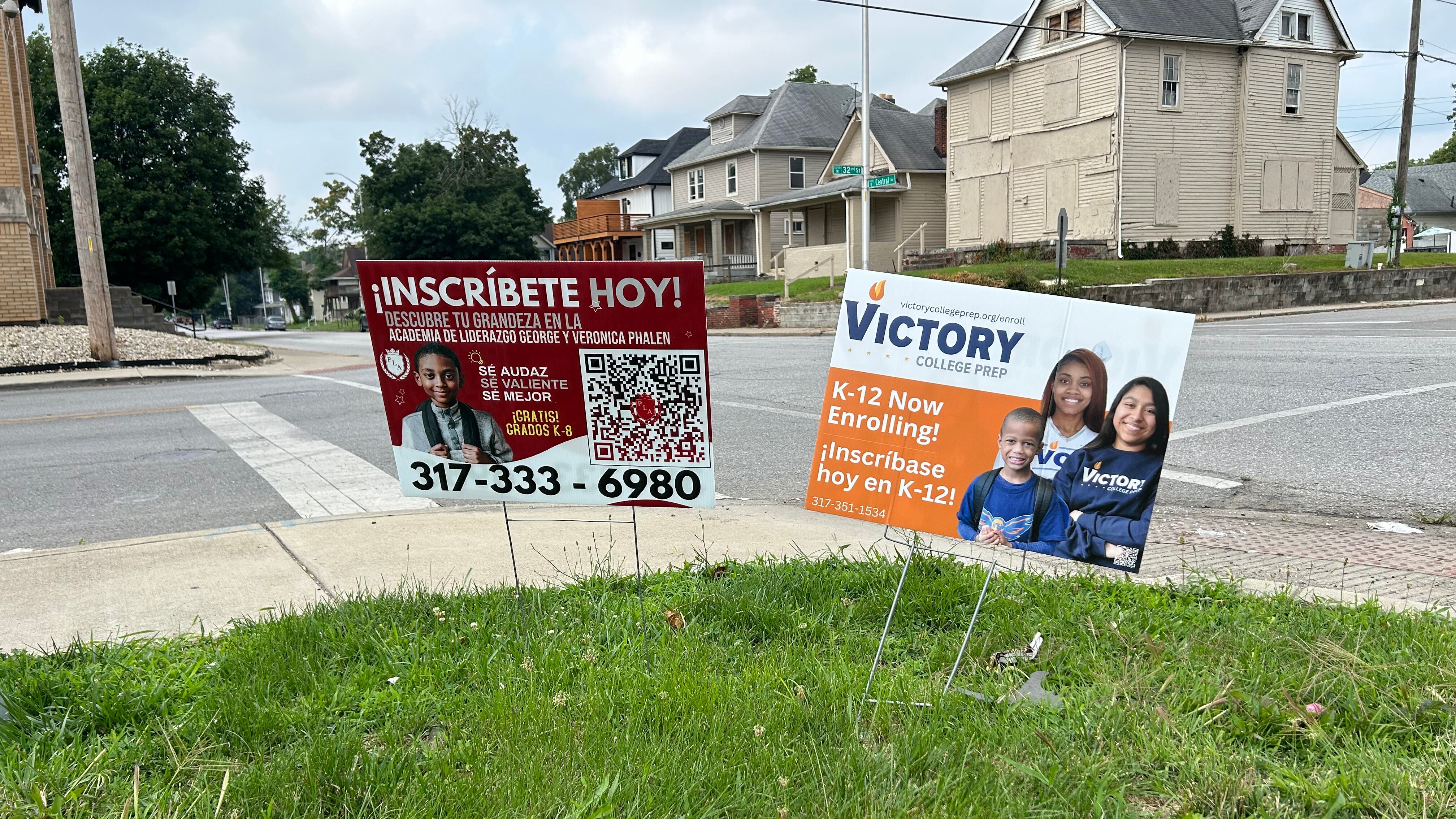 Two signs sit in grass on the side of a sidewalk. One says in Spanish, “Inscribete Hoy!” and another says “Victory College Prep.” Both signs encourage families to enroll.