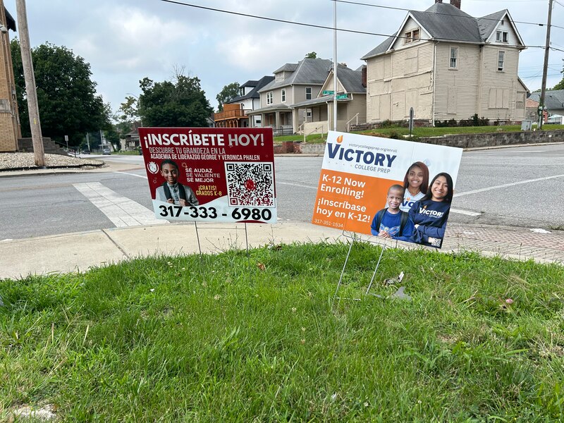 Two signs sit in grass on the side of a sidewalk. One says in Spanish, “Inscribete Hoy!” and another says “Victory College Prep.” Both signs encourage families to enroll.