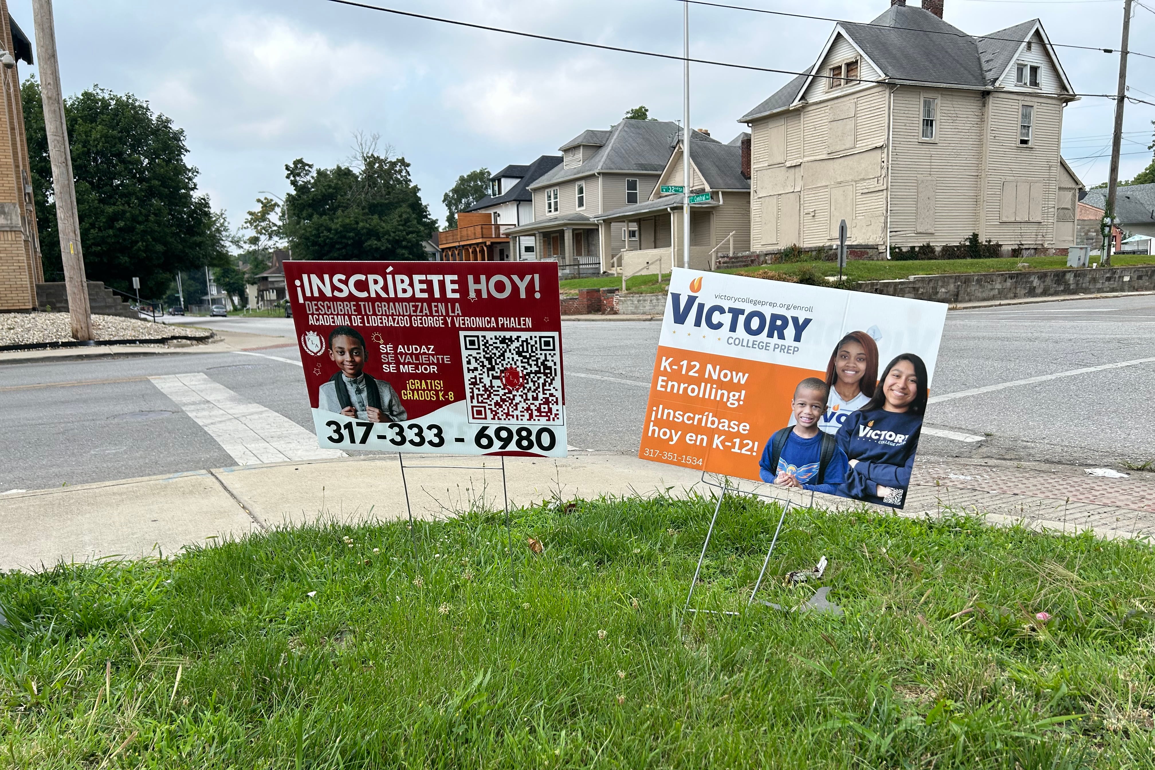 Two signs sit in grass on the side of a sidewalk. One says in Spanish, “Inscribete Hoy!” and another says “Victory College Prep.” Both signs encourage families to enroll.