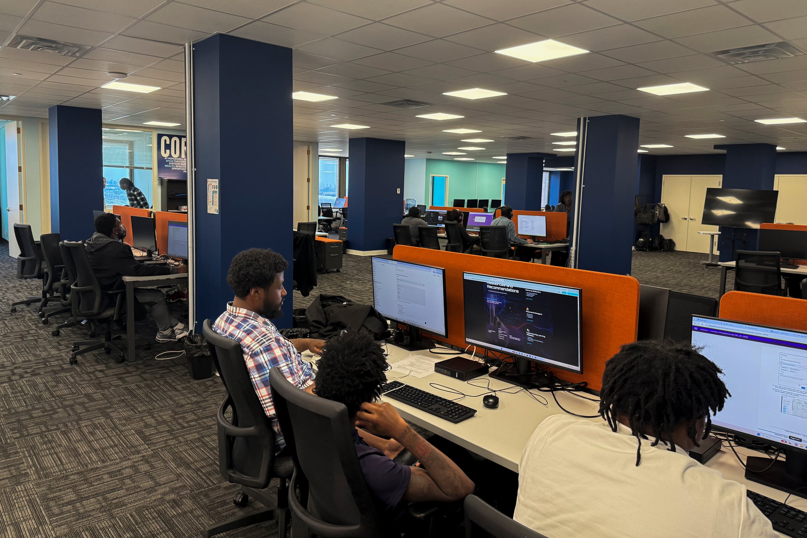 A room with people sparsely seated at workstations with a screen in front of them.