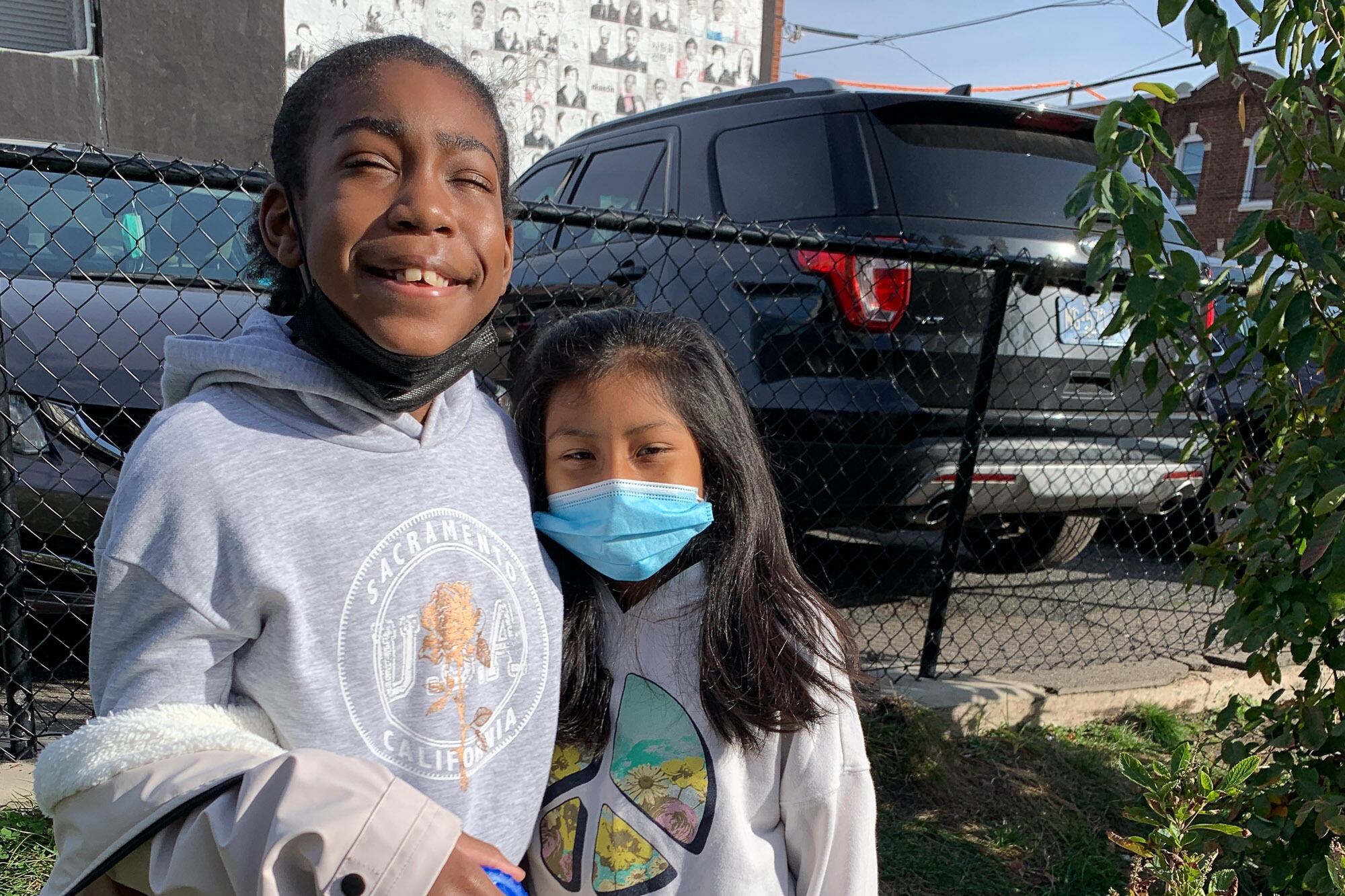 Two fourth grade students wearing hoodies pose together for a portrait in a small green space next to a parking lot.