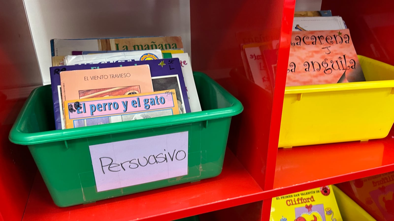 Multicolored bins sit on red shelves in a classroom. They contain Spanish language books.