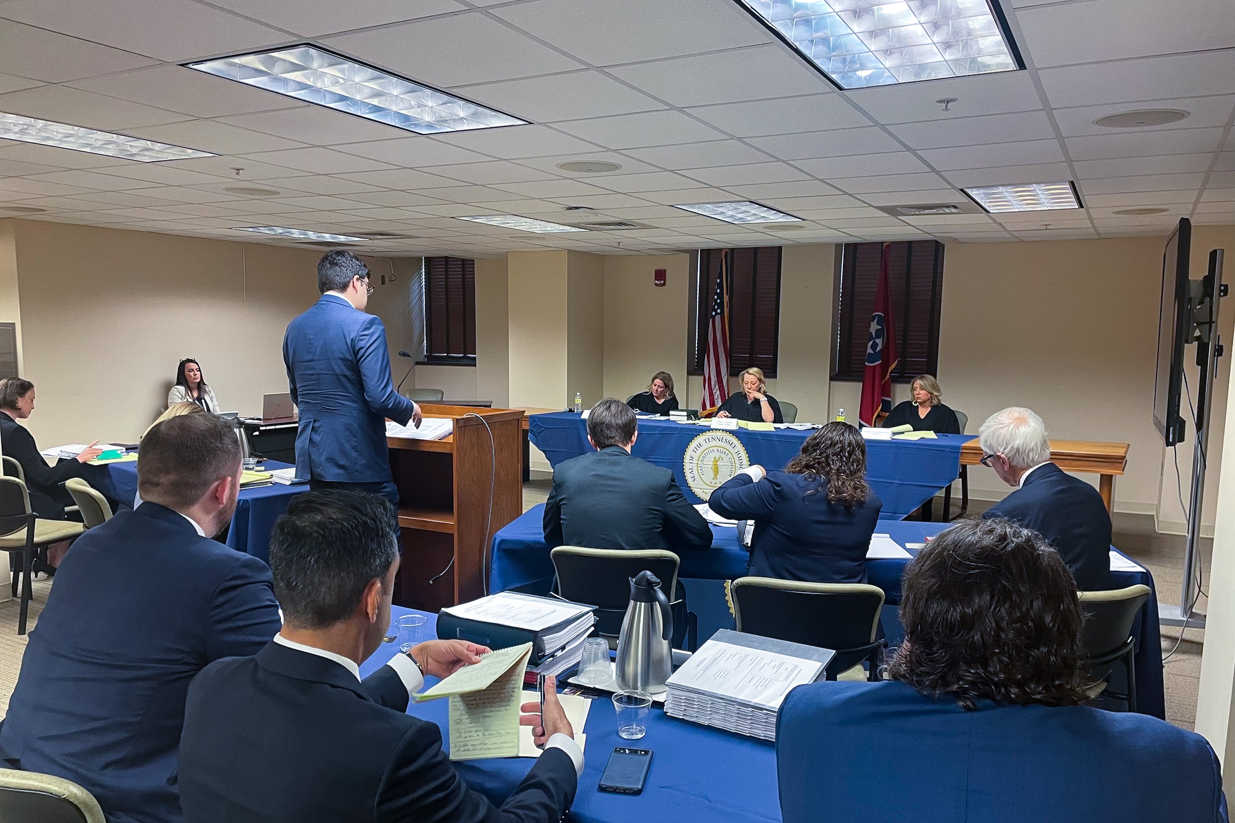 A man wearing a blue suit stands at a podium facing a judicial panel of three women wearing black robes.