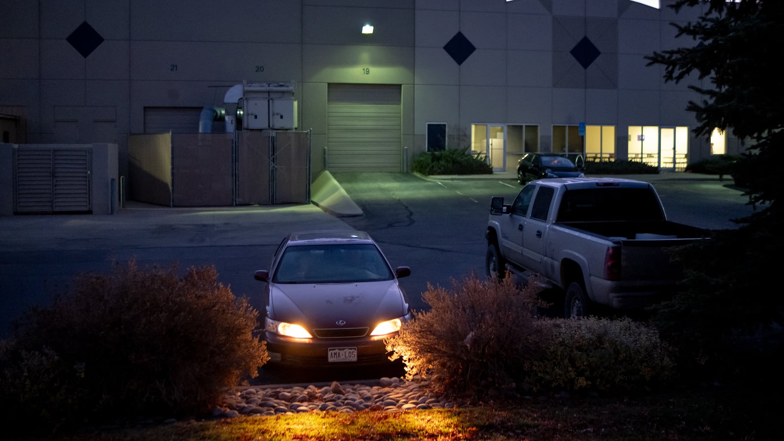A car sits with its lights on in the parking lot of a factory.
