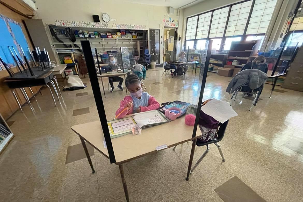 A kindergarten student sits at a desk with safety barriers inside a classroom.