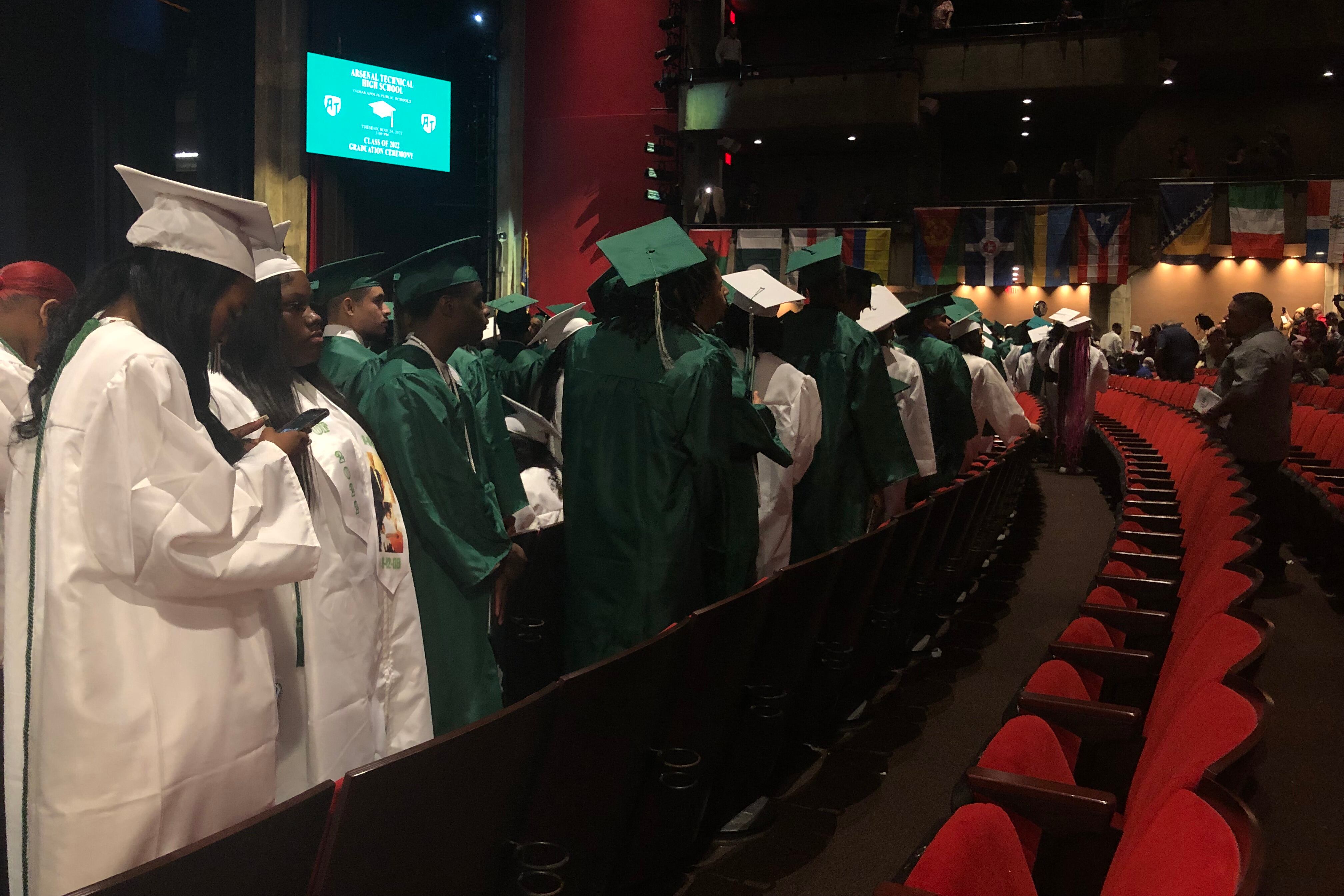 Students in both green and white graduation regalia stand in an auditorium in front of red theater seats.